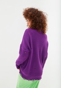 Maglione oversize viola a maglia con dettagli usurati, dotato di maniche larghe e orlo sfilacciato. Indossato sopra pantaloni verdi a trama.