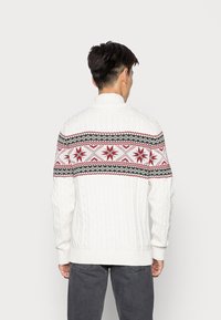 Muž otočený zády nosí bílý svetr s pleteným vzorem v červené a černé barvě přes ramena a horní část rukou, v kombinaci s tmavými džínami.