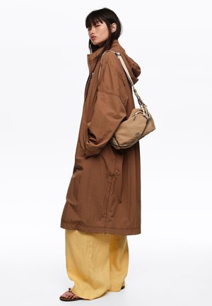 Persona joven que lleva un abrigo largo marrón y pantalones anchos amarillos, lleva un bolso beige de hombro con una correa anudada, de pie de lado sobre un fondo blanco.