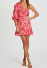 Asymetrické růžové šaty s jedním nafouknutým rukávem, s tečkovaným vzorem a řaseným lemem, vyrobené z lehkého materiálu.