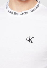 Biała koszulka z czarnym haftowanym logotypem "cK" na piersi i wzorem tekstowym "Calvin Klein Jeans" wokół kołnierza.