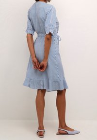 Vestido a rayas azul y blanco con mangas cortas abullonadas, cinturilla ajustable y un dobladillo con volantes. Combinado con zapatos de tacón con tira en color púrpura claro.