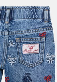 Spodnie dżinsowe w niebieskim odcieniu, z elastyczną talią i haftowanymi detalami, w tym kokardkami oraz tekstem "Guess" w różnych kolorach.