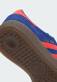 Baskets en daim bleu avec accents rouges, dotées d'une semelle extérieure en caoutchouc texturée et d'un logo doré. Le design comprend les trois bandes classiques.