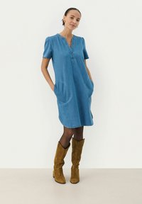 Robe en denim de couleur bleu clair, avec des manches bouffantes courtes et un décolleté en V. Présente de grandes poches et un ourlet arrondi. Portée avec des bottes hautes en daim marron.