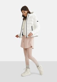 Doudoune blanche avec fermeture éclair et capuche, assortie à une jupe rose clair, des collants et des baskets blanches. Tissu texturé avec des coutures visibles.