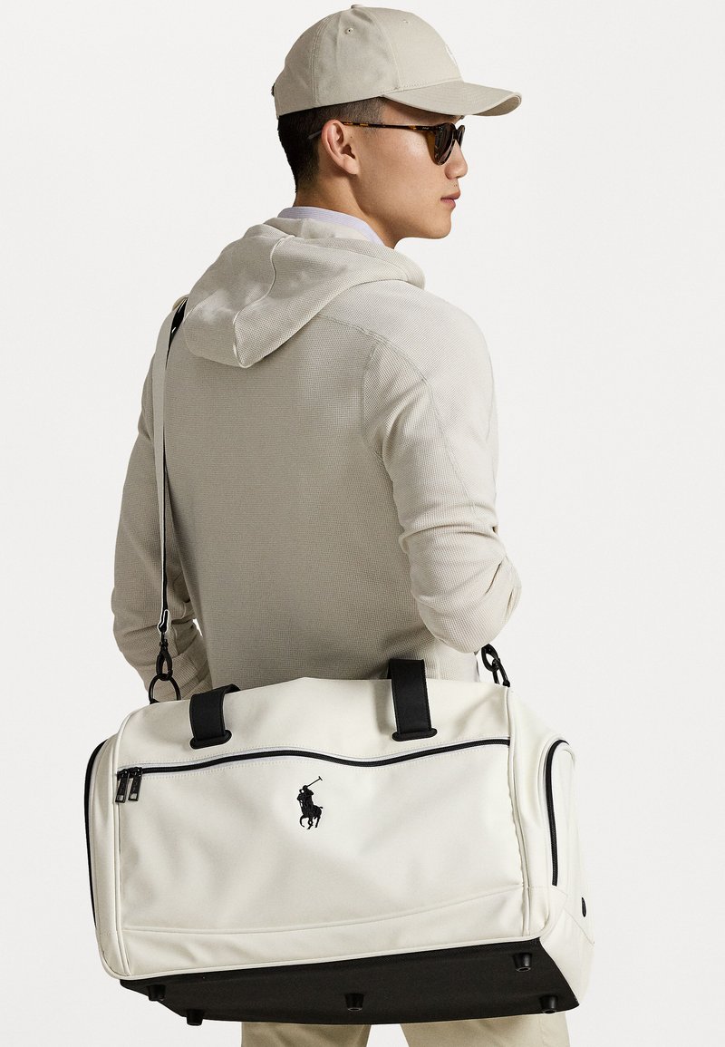 Hvid duffelbag med sorte detaljer og lynlås, der har et logo. Modellen bærer lyse tøj, en kasket og solbriller, mens han holder tasken.