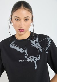 Sort bomulds T-shirt med et stort gråt sommerfugle grafik og teksten "TAKE FLIGHT." Rund hals og korte ærmer. Glat tekstur.