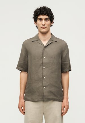 Jeune homme aux cheveux bouclés et foncés portant une chemise marron à manches courtes boutonnée et un pantalon beige clair, debout devant un fond uni.