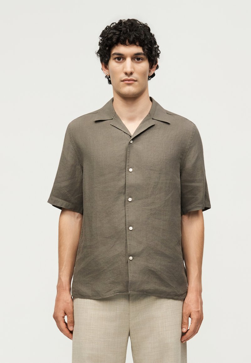 Νέος άνδρας με σγουρά σκούρα μαλλιά, φορώντας ένα καφέ κοντομάνικο πουκάμισο με κουμπιά και ανοιχτό μπεζ παντελόνι, στέκεται μπροστά σε ένα απλό φόντο.