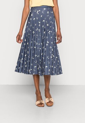 Frau trägt einen marineblauen plissierten Midirock mit weißem Blumenmuster, beige Slidesandalen und ein gelbes Oberteil, steht vor einem schlichten Hintergrund.