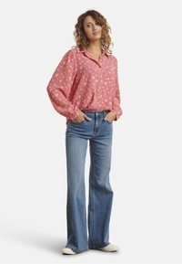 Pink long-sleeve blouse with a heart pattern, collared neckline, and elastic cuffs, paired with light blue flared jeans and white sneakers.