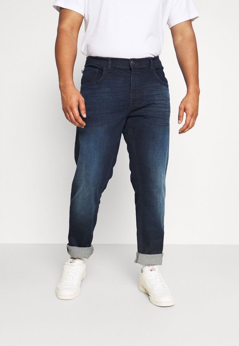 Jeans de mezclilla azul oscuro con un corte relajado, dobladillos enrollados, cinco bolsillos y detalles de costura en contraste, combinados con zapatillas blancas.