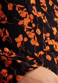 Svart tyg med en texturerad yta, med en orange blommig tryck. Inkluderar samlad detalj vid ärmslutet och en jämn drapering.