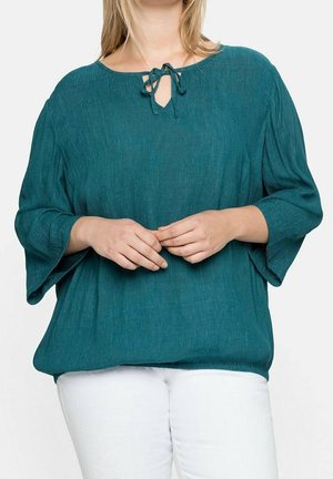 Teal textured blouse with a round neckline and tie detail, featuring wide sleeves and a relaxed fit, paired with white pants.