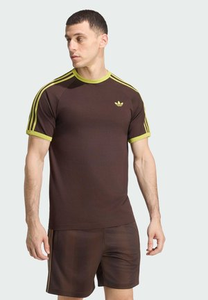 Mand iført brun Adidas t-shirt med gul kant og striber samt matchende brune shorts, stående foran en ensfarvet lys baggrund.