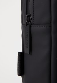 Zaino nero con una texture liscia, caratterizzato da una cerniera con un tirante contrassegnato "Calvin Klein" e una tracolla per un trasporto facile.