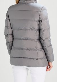 Kvinde iført en lys grå, quiltet puffjakke set fra ryggen, parret med hvide bukser mod en ensfarvet baggrund.