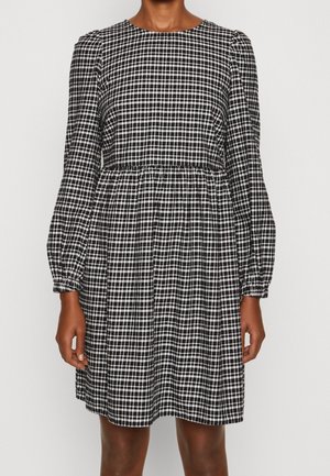 Vrouw die een zwart-wit geruite lange mouwen jurk draagt met een geplooide taille en pofmouwen, staand tegen een effen achtergrond.