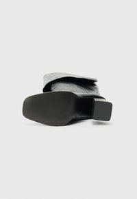 Μαύρο δερμάτινο μποτάκι που βρίσκεται στο πλάι, προβάλλοντας την υφή της σόλας και την τετράγωνη φτέρνα του σε φόντο απλό.