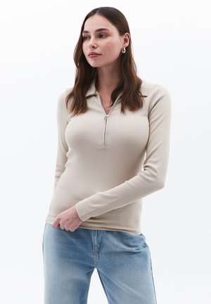 Beige geribbelde langwerpige top met een kwart-rits kraag, gecombineerd met lichtblauwe denim. De stof is glad en heeft een getailleerd ontwerp.