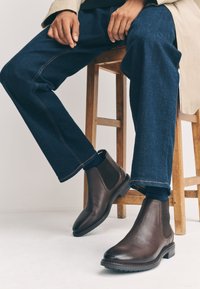 Brune Chelsea-støvler i læder med elastiske sider, gummisåler og tekstureret finish, kombineret med mørkeblå denimjeans.