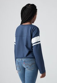 Vrouw met gevlochten haar, gekleed in een blauwe sweatshirt met witte strepen op de mouwen en blauwe jeans, met de rug naar de camera gericht tegen een effen achtergrond.