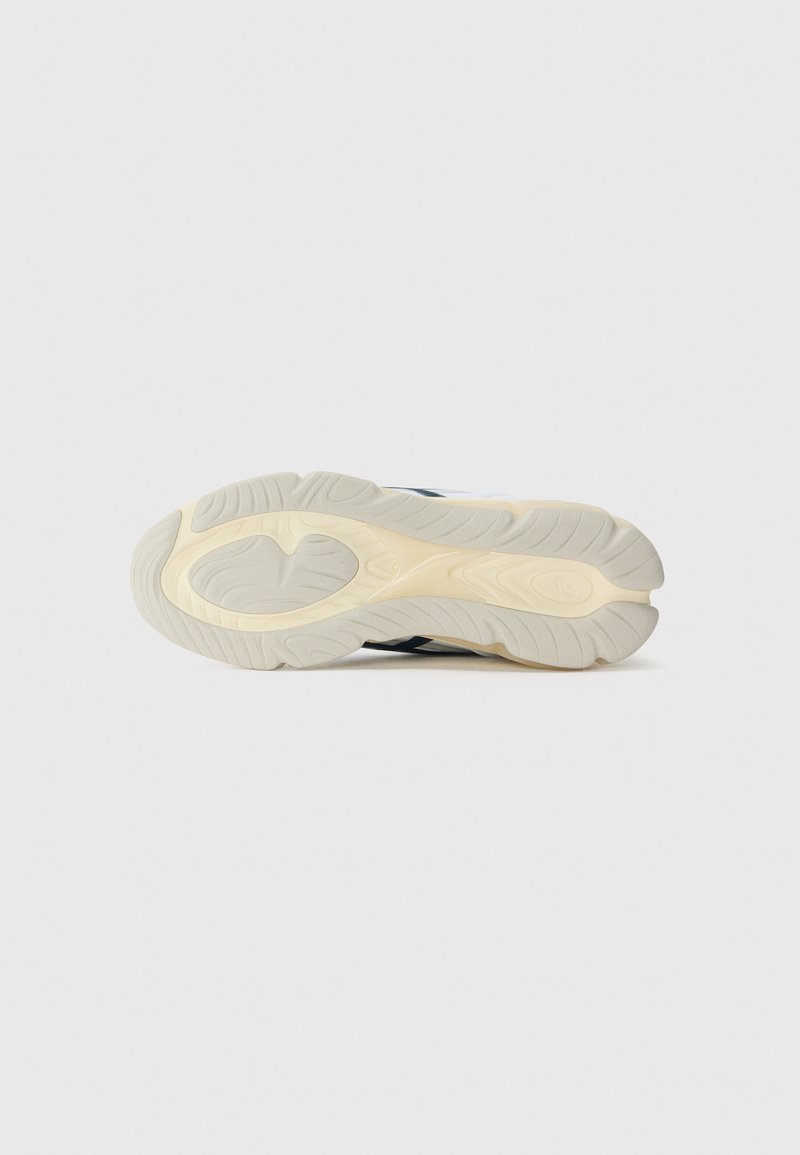 Semelle de chaussure de sport blanche avec un motif texturé et ondulé, dotée d'une semelle extérieure en caoutchouc beige et d'accents noirs sur les côtés.
