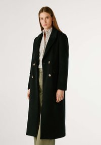 Cappotto di lana nera con design doppiopetto, bottoni argentati, rever a notched e tasche laterali. Il modello indossa una camicia a quadri sotto.