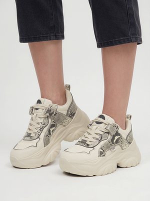 Kraftige beige sneakers med slangeprintdetaljer båret med cropped sorte bukser på en lys baggrund.