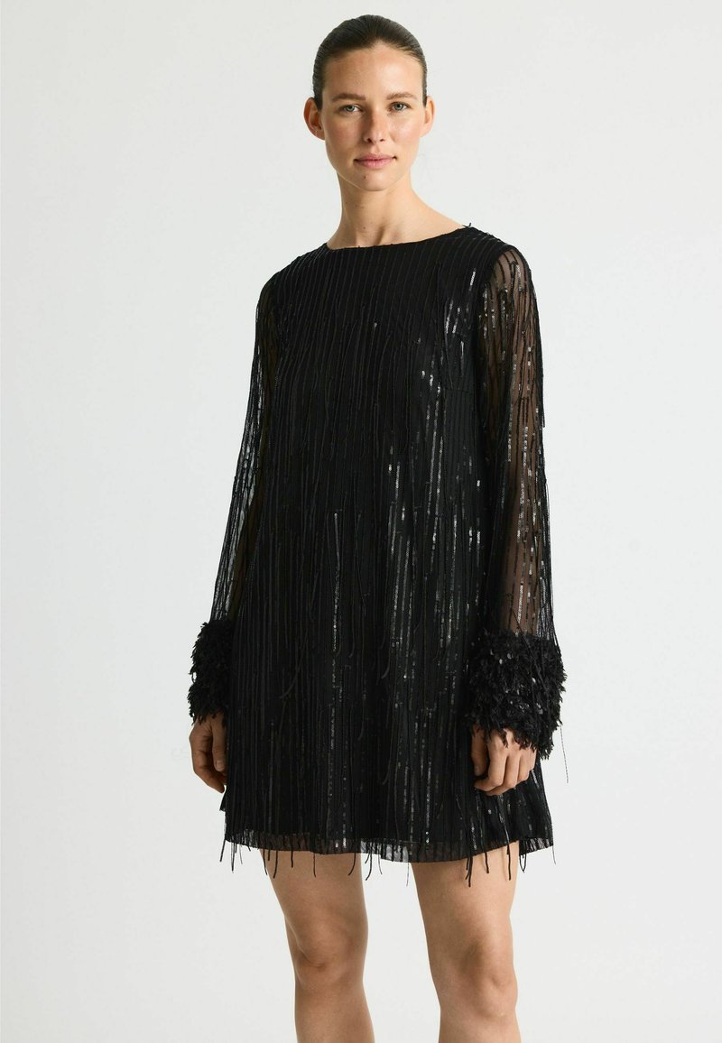 Vestito nero con maniche lunghe e sfilacciate; presenta strisce verticali di paillettes e polsini neri piumosi. La trama trasparente aggiunge profondità al design.