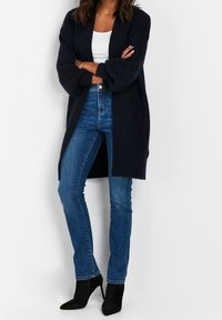 Cardigan long bleu marine en maille texturée, associé à un jean bleu taille haute et des escarpins noirs à bouts pointus. Haut blanc minimaliste en dessous.