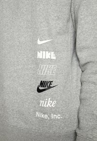 Šedá mikina s více logy Nike v různých stylech a barvách, s bílým a černým textem na strukturované tkanině.