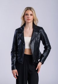 Kvinna med långt blont hår, iklädd svart läderjacka över vit crop top och lösa svarta byxor, står mot en enkel ljus bakgrund.