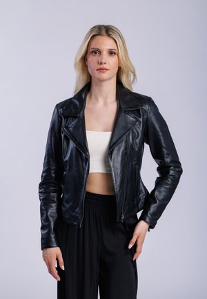 Femme aux longs cheveux blonds portant une veste en cuir noir sur un crop top blanc et un pantalon noir ample, se tenant devant un fond clair.