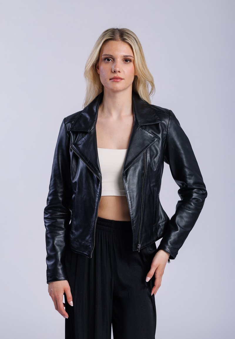 Kvinna med långt blont hår, iklädd svart läderjacka över vit crop top och lösa svarta byxor, står mot en enkel ljus bakgrund.