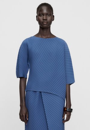 Femme portant un haut bleu texturé et une jupe assortie à plis diagonaux, debout devant un fond clair uni.