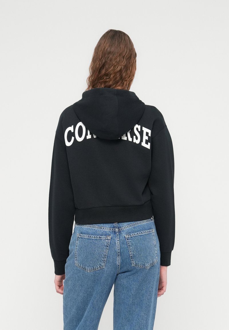 Sort crop hoodie med hvid "CONVERSE" skrift på ryggen, kombineret med lyseblå denimbukser med baglommer.