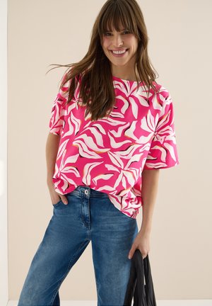 Femme souriante aux longs cheveux bruns portant un haut à motifs roses et blancs et un jean bleu, tenant une veste noire dans une main.