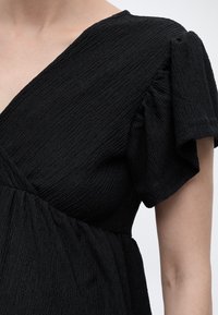 Femeie purtând o rochie neagră din material texturat, cu decolteu în V și mâneci scurte evazate, prezentată de la umăr până la talie.