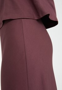 Viininpunainen vaate, jossa on väljä yläosa, joka peittää tiukan alaosan. Kangas näyttää sileältä, ja siinä on pieni kiilto. Ei näkyviä kuvioita tai yksityiskohtia.