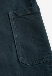 Donkerdenimstof heeft een zak met een angulaire vorm en contrasterende stiksels. De textuur lijkt iets ruw met een subtiele glans.
