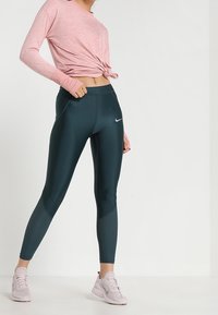 Hellrosa, langärmliges Oberteil, das an der Taille geknotet ist, kombiniert mit dunkelblauen Leggings mit einem strukturierten Muster und einem kleinen Logo, getragen mit hellrosa Sneakers.