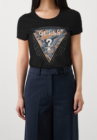 Svart t-shirt med kort ärm som har en grafisk triangellogo i krämfärg och beige, prydd med strass för accent. Matchas med marinblå byxor.