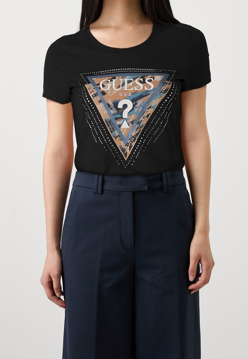 Svart t-shirt med kort ärm som har en grafisk triangellogo i krämfärg och beige, prydd med strass för accent. Matchas med marinblå byxor.