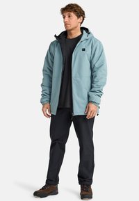 Chaqueta impermeable de color azul claro con capucha, puños elásticos y cremallera frontal. Combinada con pantalones negros y botas de senderismo marrones.