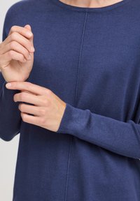 Pulover bleumarin cu manșete elasticizate și decolteu rotund; prezintă un detaliu cu cusătura verticală pe față. Textura moale, tricotată este vizibilă.