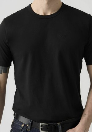 Mann iført enkel svart t-skjorte med rund hals, mørke jeans og svart belte, med tatoverte armer mot en ensfarget lys bakgrunn.