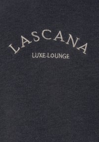 Donkergrijze stof met ribbelstructuur, voorzien van een geborduurde "LASCANA" in het wit in een boog boven de tekst "LUXE-LOUNGE" in het midden.