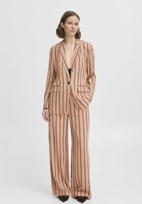 Completo a righe rosa chiaro con pantaloni a gamba larga e blazer su misura. Presenta una scollatura profonda e due tasche frontali. Tessuto testurizzato con linee verticali.